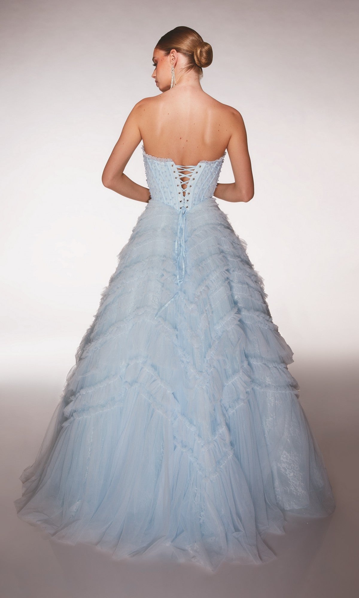 A woman poses with her back to the camera, wearing the Alyce 62208 Long Prom Dress—a strapless light blue ballgown with a lace-up corset and layered tulle ruffle skirt.